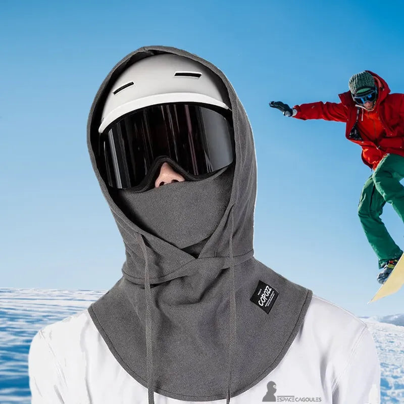 Cagoule par dessus casque ski utilisée en montagne, portée avec un casque blanc et masque de ski, snowboarder en arrière-plan sur ciel bleu.