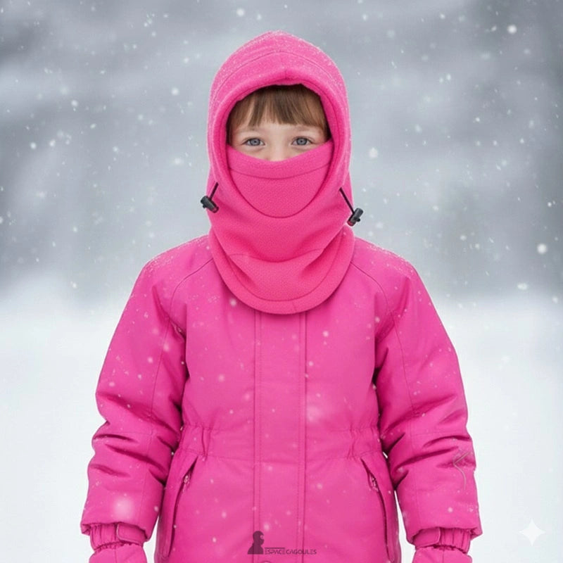 Cagoule polaire enfant rose couvrante portée par une fillette en combinaison d’hiver, debout sous la neige avec fond de forêt flou et flocons en suspension