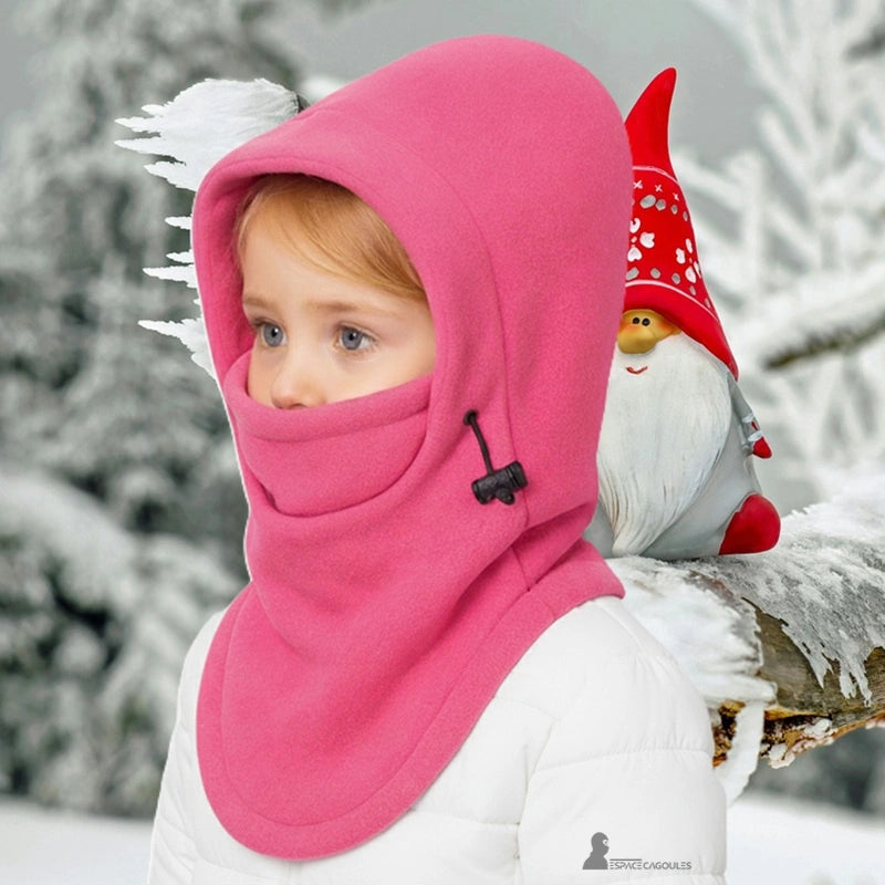 Cagoule polaire enfant rose portée par une petite fille en manteau blanc, debout dans une forêt enneigée avec un nain rouge décoratif posé sur une branche