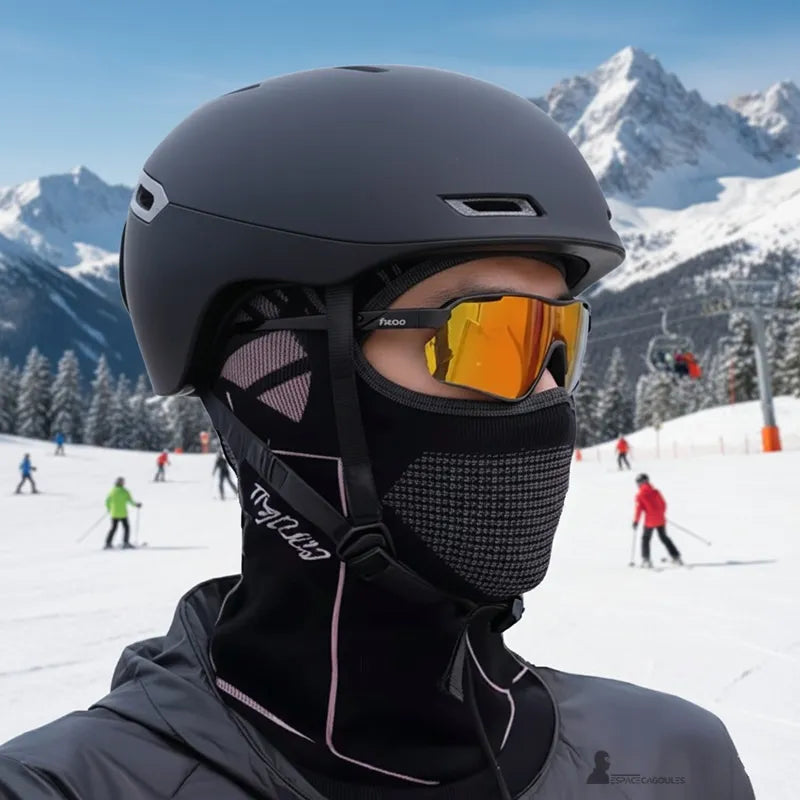 Schwarze Skimütze für Männer, getragen mit Helm und Skimaske auf einer verschneiten Piste, Dreiviertelansicht mit Bergen und Skifahrern im Hintergrund - Cagoules Bereich