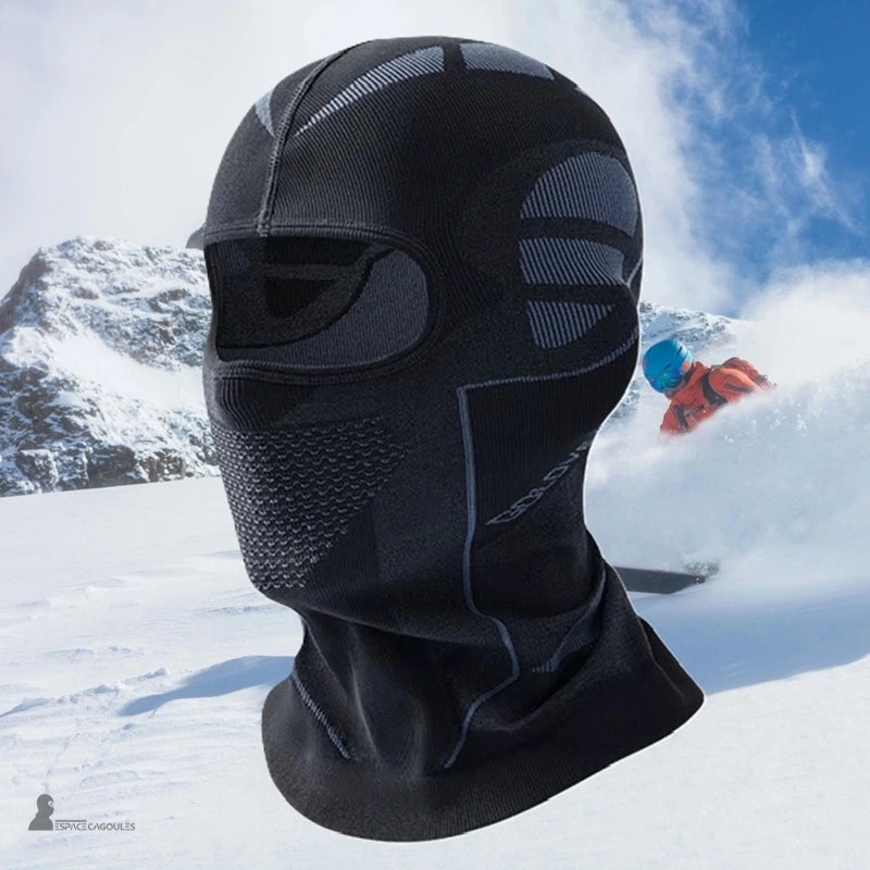 Cagoule ski homme noire technique portée en montagne, protection intégrale visage avec skieur en arrière-plan sur piste enneigée