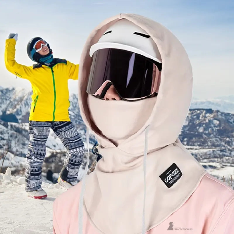 Cagoule sur casque de ski crème portée par-dessus un casque blanc avec masque, skieuse en arrière-plan sur fond de montagne enneigée - Espace Cagoules