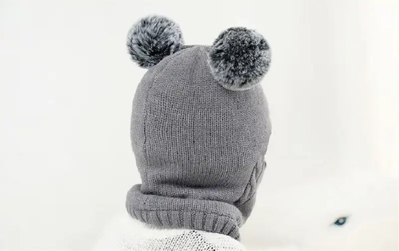 Vue arrière d’une cagoule tricot enfant grise avec deux pompons moelleux. La texture épaisse et chaude protège la tête et le cou du froid.