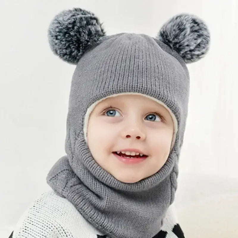Petit enfant vêtu d’une cagoule tricot enfant grise avec deux pompons sur le dessus. La matière douce et chaude garde la tête et le cou bien couverts, parfait pour les journées froides.