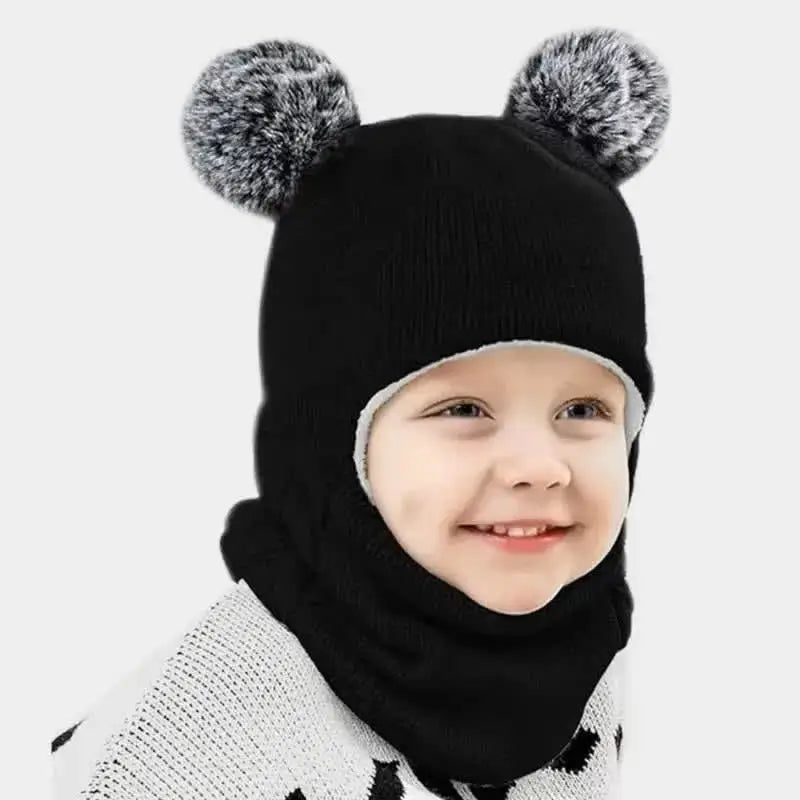 Petit garçon souriant avec une cagoule tricot enfant noire et des pompons gris. Ce modèle protège parfaitement du froid avec une touche originale.