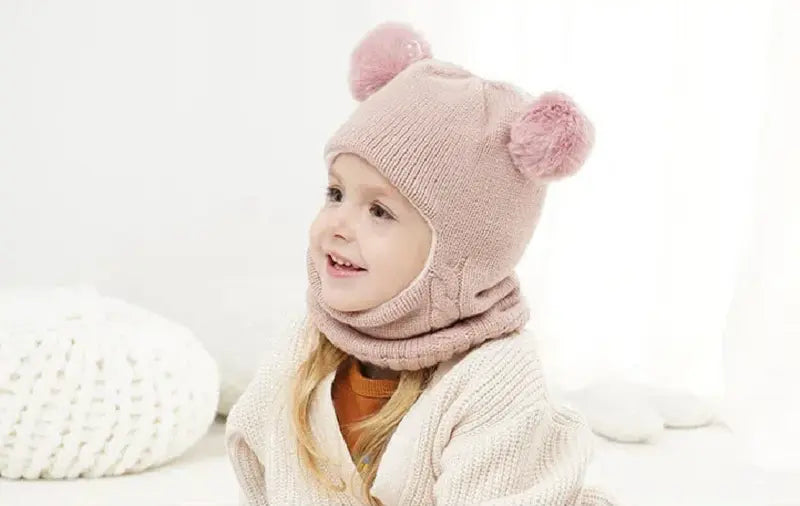 Petite fille souriante portant une cagoule tricot enfant rose avec deux pompons sur le dessus. La cagoule couvre la tête et le cou, offrant une protection douce et chaude contre le froid.