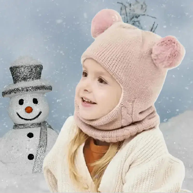 Enfant portant une cagoule tricot enfant rose avec deux pompons en forme d'oreilles, debout dans un paysage enneigé avec un bonhomme de neige en arrière-plan. La cagoule couvre bien la tête et le cou pour protéger du froid.