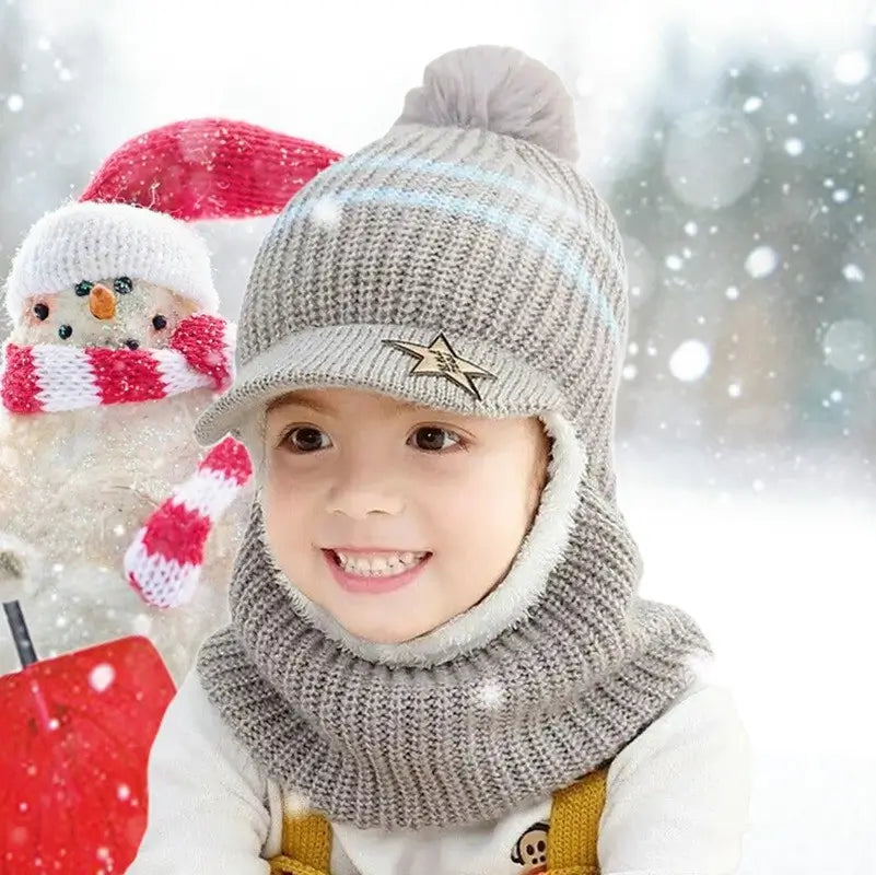 Enfant souriant portant une casquette bonnet enfant en laine grise tricotée avec pompon, visière et doublure douce, assis devant un bonhomme de neige avec écharpe rouge et blanche, dans un décor enneigé hivernal.