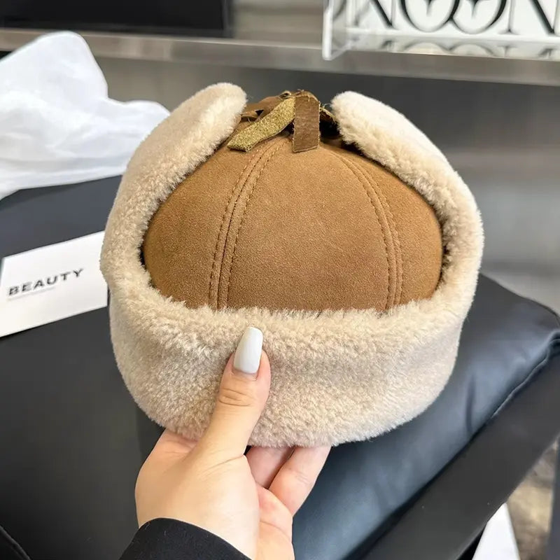 Casquette Chapka beige en cuir avec une bordure épaisse en fausse fourrure beige. La main d’une personne avec des ongles manucurés tient la casquette, mettant en valeur ses finitions douces et son style rétro chic.