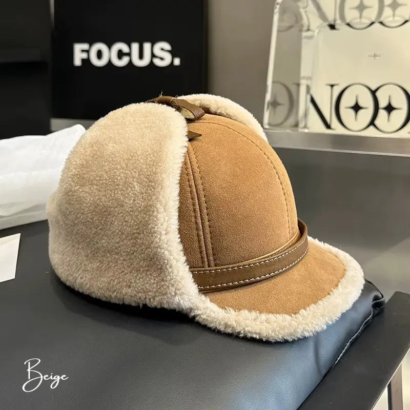 Casquette Chapka beige avec une bordure en fausse fourrure beige. Son design élégant avec oreilles rabattables en fait un accessoire idéal pour l’hiver. La casquette est posée sur un canapé en cuir avec un fond moderne.