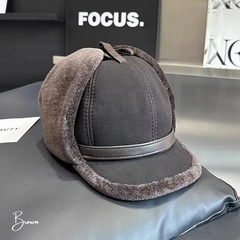 Casquette Chapka brown avec rabats en fausse fourrure grise et un petit nœud décoratif sur le dessus. Parfaite pour un look hivernal à la fois moderne et classique. La casquette est posée sur un siège en cuir avec un fond sobre.