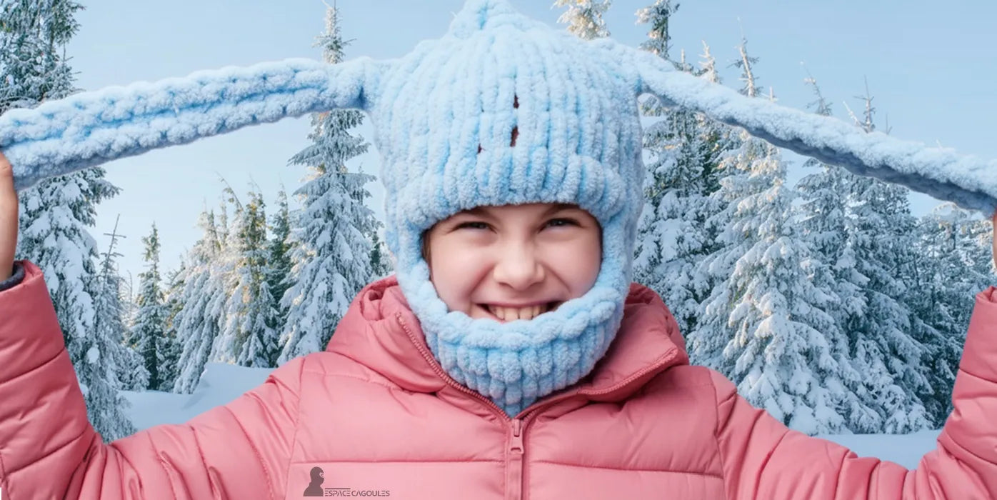 Cagoule enfant bleue en laine épaisse avec oreilles, portée par une fillette souriante en manteau rose dans une forêt enneigée – Cagoule Enfant Hiver - Espace Cagoules