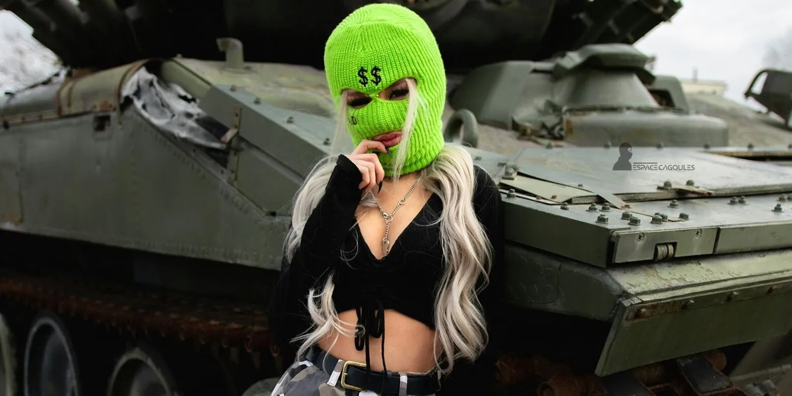 Cagoule fluo verte avec symbole dollar, femme devant un char militaire, contraste entre mode urbaine et décor industriel