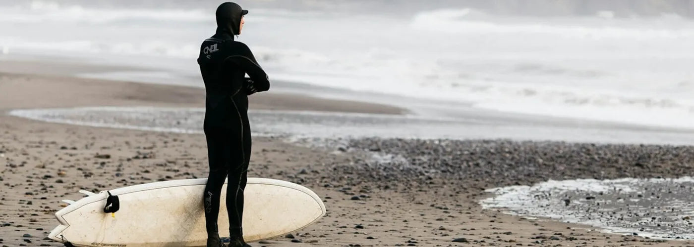 Surfeur équipé d’une combinaison et d’une cagoule néoprène, observant les vagues sur une plage déserte – Cagoule de Surf Espace Cagoules