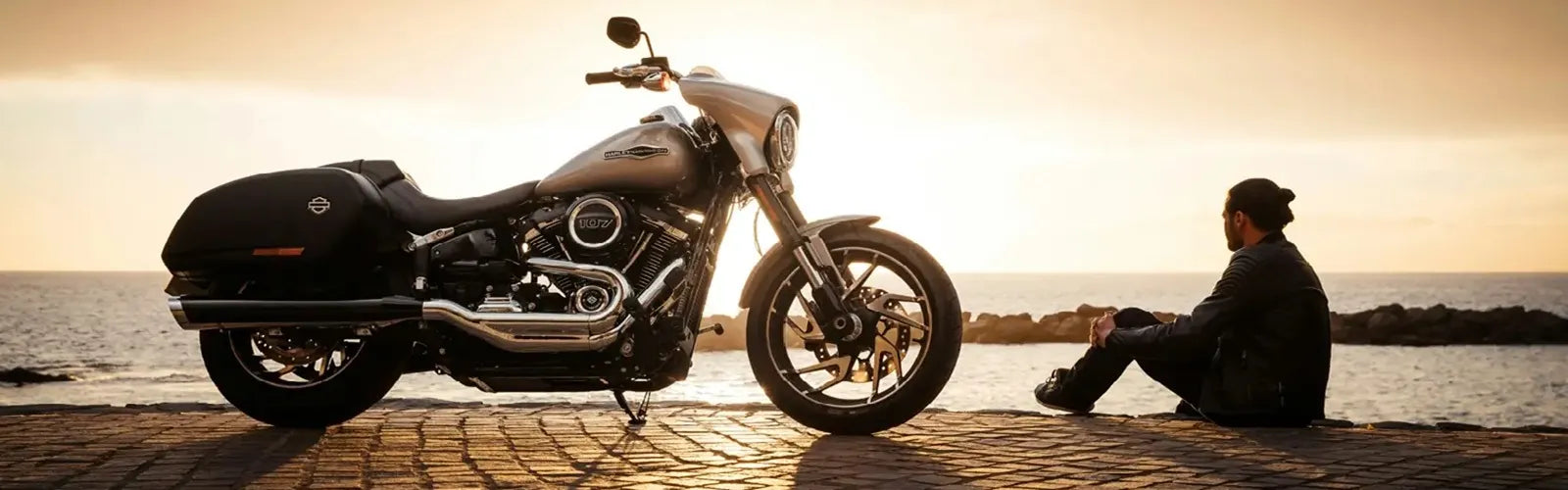 Motard assis face à sa moto sur une route côtière au coucher du soleil, représentant la collection Cagoules Moto – Espace Cagoules