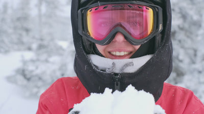 Femme souriante en tenue de ski complète, soufflant sur sur la neige détenue dans ses mains.